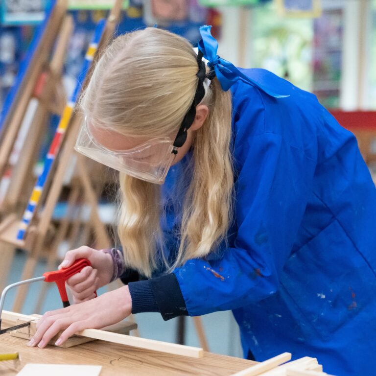 Person in a blue smock and safety goggles leans over a wooden workbench, using a red-handled saw to cut a piece of wood in a classroom workshop.
