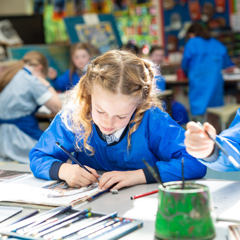 Girl with braided hair in a blue art smock sketching at a classroom table, with paints and brushes nearby as classmates work in the background.