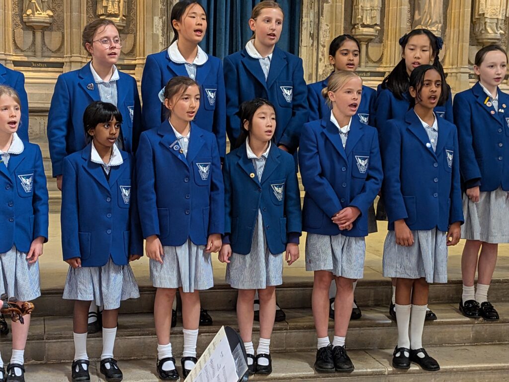 Group of schoolgirls in blue blazers singing on stone steps indoors.