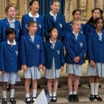 Group of schoolgirls in blue blazers singing on stone steps indoors.