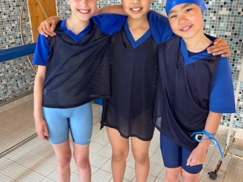 Three kids wearing blue swim caps and black training vests stand arm in arm by a tiled pool area, smiling at the camera.