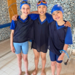 Three kids wearing blue swim caps and black training vests stand arm in arm by a tiled pool area, smiling at the camera.