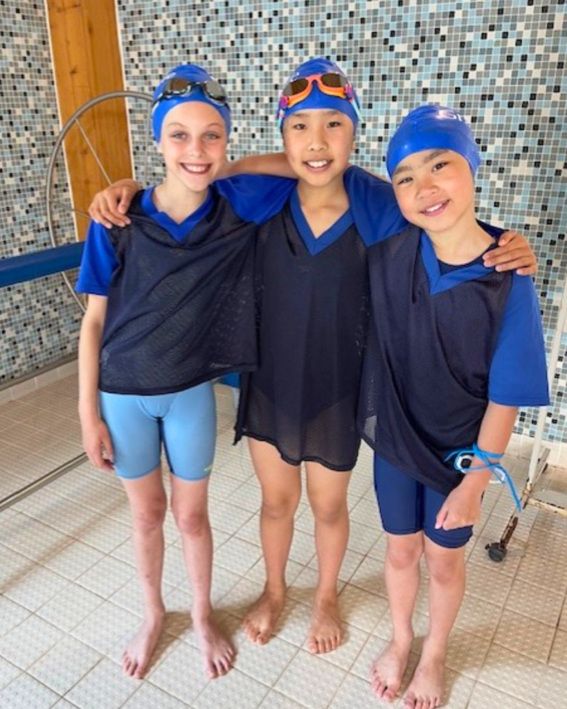 Three kids wearing blue swim caps and black training vests stand arm in arm by a tiled pool area, smiling at the camera.